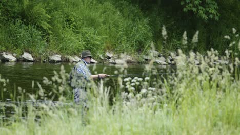 Un-Hombre-Con-Sombrero-Y-Gafas-De-Sol-Se-Encuentra-En-Un-Río,-Pescando-Con-Mosca