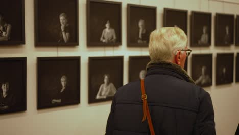 Una-Persona-De-Pelo-Gris-Mira-Una-Fila-De-Fotografías-En-Blanco-Y-Negro-En-Una-Galería-De-Arte