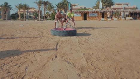 Un-Grupo-De-Atletas-Arrastra-Un-Enorme-Neumático-De-Coche-Por-La-Playa---Frente---Cámara-Lenta