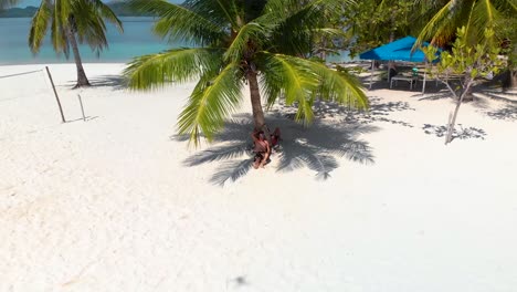 Aerial-drone-shot-away-from-a-man-sitting-under-a-palm-tree,-revealing-a-tropical,-paradise-isle-and-Paraw-boats,-in-turquoise-sea,-at-Pass-island,-in-Philippines,-Asia