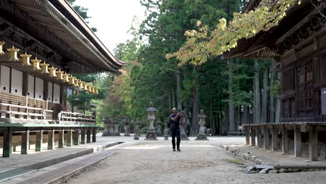 Solo-Male-Wearing-Backpack-Walking-Along-Path-Looking-At-Row-Of-Hanging-Golden-Lanterns-At-Koyasan,-Taking-Photos-Using-Mobile-Phone