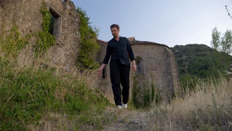 Toma-Baja-En-Cámara-Lenta-De-Un-Hombre-Vestido-De-Negro-Caminando-Sobre-Un-Campo-De-Hierba-Afuera-De-Las-Ruinas-De-Un-Castillo-Durante-La-Puesta-De-Sol