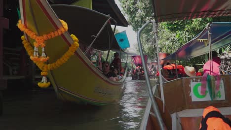 Toma-De-Establecimiento,-Bote-Con-Turistas-Pasando-Y-Saludando-Con-El-Signo-De-La-Paz-En-El-Mercado-Flotante-De-Tailandia,-Bote-Bomba-Alineado-En-El-Fondo