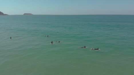 Drone-shot-of-people-surfing-in-the-clear-and-crystal-ocean-of-the-Santinho-Beach,-Brazil,-waiting-for-the-waves,-in-their-surfing-wetsuits-on-a-sunny-day