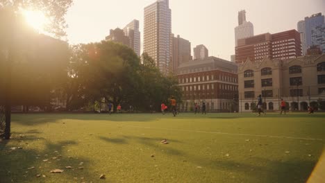 Weitwinkelaufnahme-Von-Zwei-Mannschaften,-Die-Bei-Sonnenuntergang-Auf-Einem-Rasenplatz,-Umgeben-Von-Einem-Wolkenkratzer,-In-New-York-Fußball-Spielen