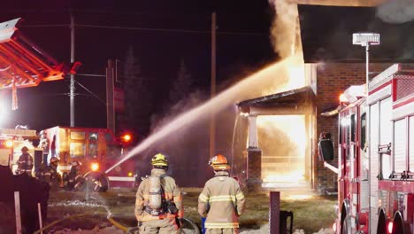 Feuerwehrleute-Schließen-Den-Schlauch-An-Und-Sprühen-In-Richtung-Vorderseite-Des-Hauses,-Um-Nachts-Das-Feuer-Zu-Löschen