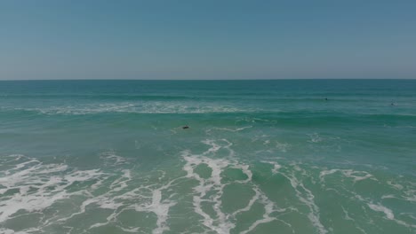 Toma-De-Drone-De-Personas-Surfeando-En-El-Océano-Claro-Y-Cristalino-De-La-Playa-De-Santinho,-Brasil,-Esperando-Las-Olas,-En-Sus-Trajes-De-Surf-En-Un-Día-Soleado