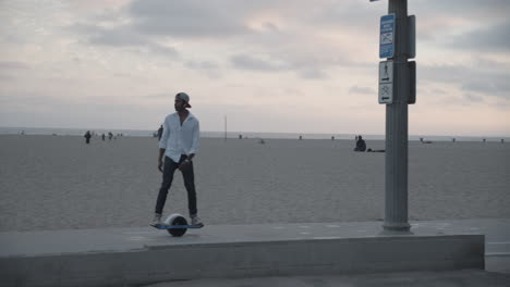 Toma-En-Cámara-Lenta-De-Un-Hombre-Montando-Patineta-Electrónica-Con-La-Playa-Al-Fondo-Durante-La-Puesta-De-Sol-En-Santa-Mónica,-Ca