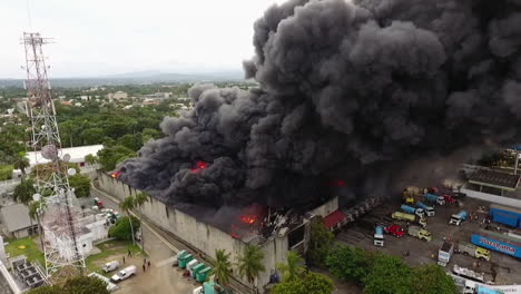 Vista-Aérea-De-Camiones-De-Bomberos-Y-Bomberos-Luchando-Contra-Un-Almacén-En-Llamas,-Humo-Negro-Elevándose,-Durante-El-Día,-En-República-Dominicana---Retroceso,-Disparo-De-Drones