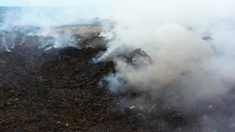 Aerial-view-of-the-firemen-using-a-Excavator-to-extinguish-a-Junkyard-fire,-smoke-rising-while-trash-burn,-cloudy-day,-in-Dominican-Republic---static,-drone-shot