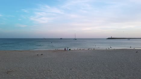 Couple-sitting-on-sandy-beach-and-staring-at-the-Atlantic-Ocean-amongst-seagulls-after-sunset-at-Sesimbra,-Portugal