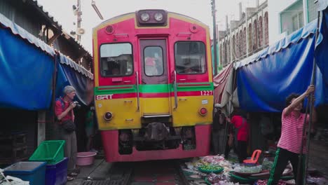 Toma-En-Primera-Persona,-El-Tren-Se-Mueve-A-Lo-Largo-Del-Ferrocarril-Del-Mercado-De-Maeklong-En-Tailandia,-Un-Turista-Toma-Una-Fotografía,-Los-Vendedores-Preparan-Sus-Tiendas-En-El-Fondo