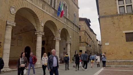 Torre-Del-Reloj-En-El-Palazzo-Comunale-En-Piazza-Pio-II-Con-Lugareños-En-La-Esquina-De-La-Calle-En-Pienza,-Toscana,-Italia