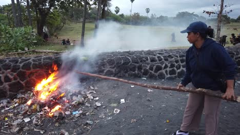 Una-Mujer-Con-Un-Palo-Quema-Basura-Plástica-Creando-Humo-Y-Fuego,-Playa-Asiática-En-Bali-Indonesia,-Humos-Tóxicos,-Contaminación