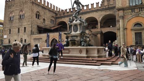 Turistas-Caminando-Y-Tomándose-Selfies-En-La-Fuente-De-Neptuno-Con-El-Palacio-Re-Enzo-Al-Fondo-En-Bolonia,-Italia