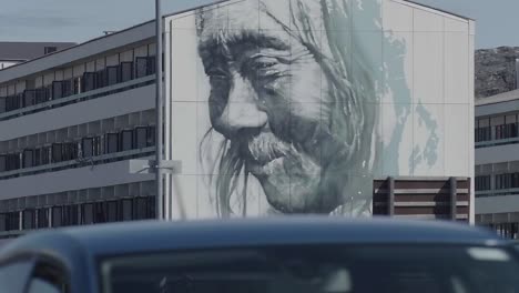 Vista-Bloqueada-Del-Artista-Guido-Van-Helten,-Arte-Mural-En-El-Costado-Del-Edificio-Con-Tráfico-Que-Pasa-En-Nuuk.