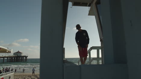 Toma-Cinematográfica-De-Un-Salvavidas-Caminando-Hacia-La-Torre-De-Vigilancia-En-La-Playa-Con-El-Histórico-Muelle-De-La-Playa-De-Manhattan-Y-Turistas-Al-Fondo