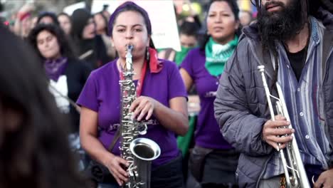 Mujeres-Vestidas-Con-Camisetas-Moradas-Tocan-El-Saxofón-Y-La-Batería-Durante-Una-Protesta-Y-Marcha-En-El-Día-Internacional-De-La-Mujer