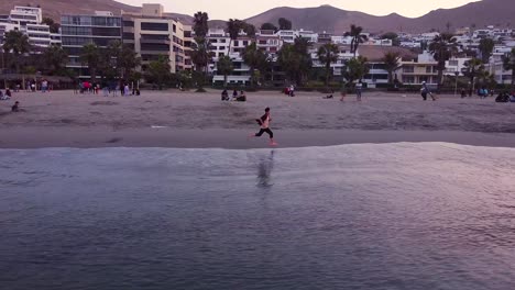 Young-boy-running-barefoot-on-a-beach-full-of-families-during-sunset