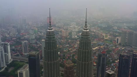 Drones-Aéreos-Vuelan-Pedestal-Hacia-Abajo-A-Lo-Largo-Del-Hito-Icónico,-Torres-Gemelas-Petronas-Klcc,-Capturando-El-Paisaje-Urbano-Del-Centro-En-El-Distrito-Central,-Kuala-Lumpur-En-Un-Día-Brumoso