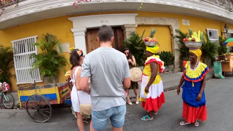 A-tall-man-and-several-women-are-standing-and-talking-to-palenqueras-with-colorful-dresses-who-are-balancing-fruit-bowls-over-their-heads