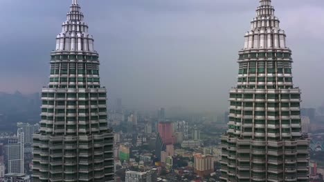 Drone-Cinematográfico-Vuela-Pedestal-A-Lo-Largo-Del-Hito-Icónico,-Torres-Gemelas-Petronas-Klcc,-Capturando-El-Paisaje-Urbano-Del-Centro-En-El-Distrito-Central,-Kuala-Lumpur-En-Un-Día-Brumoso