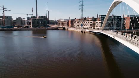 Vista-Aérea-De-Un-Barco-Conduciendo-Bajo-Un-Puente-Blanco,-Mientras-La-Gente-Camina-Sobre-él,-Día-Soleado,-En-El-área-De-Kalasatama,-Ciudad-De-Helsinki,-Uusimaa,-Finlandia---Dolly,-Disparo-De-Drone