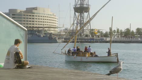 Un-Joven-Rubio-Tocando-La-Guitarra-Durante-La-Puesta-De-Sol-Junto-Al-Agua-En-El-Puerto-Comercial-De-Barcelona,-España