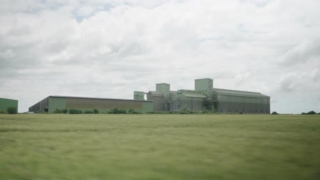 Driving-Past-Green-Wheat-Field-With-Wheat-Factory-In-The-Distance