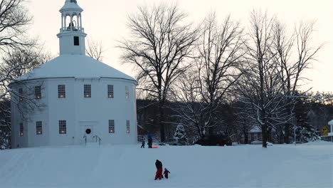 Lapso-De-Tiempo-De-Niños-Y-Adultos-En-Trineo-Bajo-El-Resplandor-Del-Atardecer-En-Invierno-Junto-A-La-Antigua-Iglesia-Redonda-En-Richmond,-Vermont