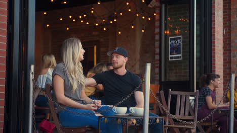 Weitwinkelaufnahme-Eines-Paares.-Ein-Mann-Blickt-Auf-Die-Uhr,-Während-Er-Mit-Einer-Jungen-Blonden-Frau-Auf-Der-Terrasse-Eines-Cafés-Spricht