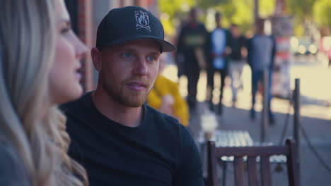 Closeup-Shot-of-Young-Man-talking-to-Young-Blonde-Woman-at-Outdoor-Café-Patio-on-Busy-Street