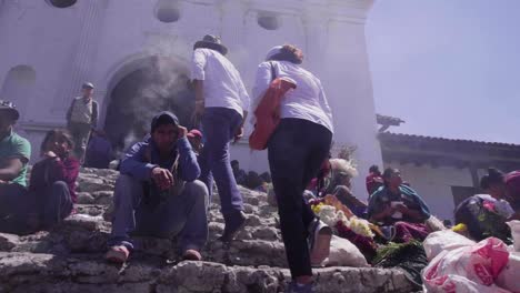 A-couple-walking-up-the-entrance-of-an-old-catholic-church-in-Chichicastenango,-Guatemala,-between-smoke-and-flowers