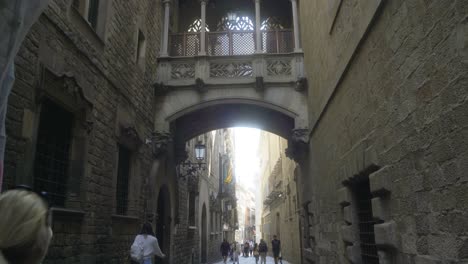 Pont-del-Bisbe,-Barcelona,-with-couple-and-tourists-walking-through-narrow-street-in-Barri-Gothic-Quarter