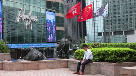 Un-Hombre-Descansa-Fuera-De-La-Plaza-De-Cambio,-El-Edificio-Que-Alberga-El-Mercado-De-Valores,-Mientras-Las-Banderas-De-La-República-Popular-China-Y-De-La-RAE-De-Hong-Kong-Ondean-Al-Viento-En-El-Fondo.