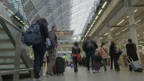 Static-shot-of-two-waiting-travellers-looking-on-as-many-people-walk-past-in-a-busy-St-Pancras-International-station-in-London