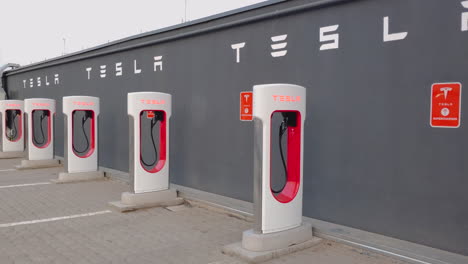 Estaciones-De-Carga-De-Supercargadores-De-Automóviles-Eléctricos-Tesla,-Sin-Gente,-Cerca-De-Una-Pared-Gris-En-El-Estacionamiento-En-La-Terraza-De-Un-Centro-Comercial-En-Rumania---Vista-Panorámica-Cercana-Del-Día-A-La-Izquierda