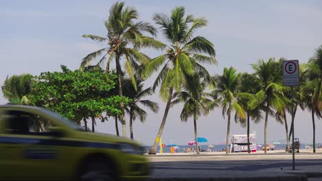 Toma-Amplia-Que-Muestra-Gente-Caminando-A-Lo-Largo-De-La-Carretera-Frente-A-Palmeras-Y-La-Playa-En-Río-De-Janeiro:-Tráfico-En-Primer-Plano.