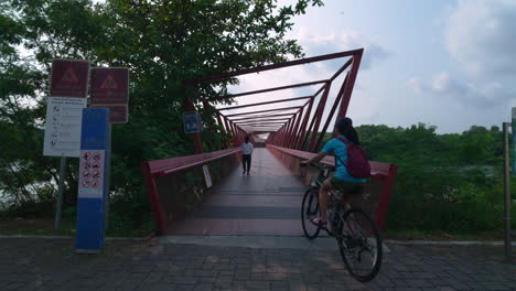 Ciclistas-Cruzando-El-Puente-Rojo-En-Cámara-Lenta-En-El-Parque-Punggol-Promenade,-Singapur