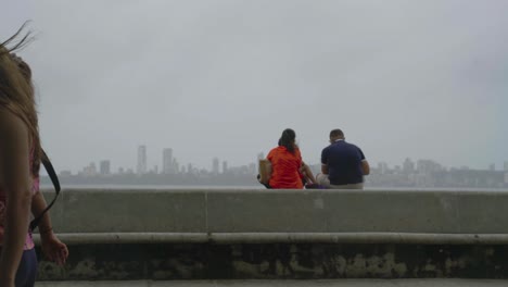 Pareja-Sentada-En-El-Terraplén-Y-Observando-La-Bahía-Trasera-Y-El-Paisaje-Urbano-Durante-La-Pandemia-Del-Coronavirus-En-Mumbai,-India