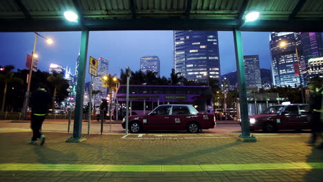 Gente-Caminando-Por-La-Acera-Cerca-De-La-Bahía-De-Taxis-En-Hong-Kong-Con-Edificios-Altos-Iluminados-Al-Fondo