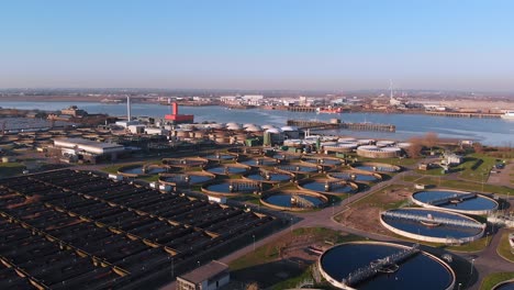 Aerial-over-Crossness-Sewage-Treatment-Works,-with-the-Thames-River-and-Boats-in-the-background