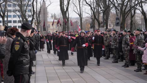 Vista-Estática-En-Cámara-Lenta-De-Veteranos-Marchando-En-Uniforme-Retirado-Por-Una-Columna-A-Través-De-Una-Multitud-Con-Policías-Y-Jóvenes-Soldados-Alineados-En-El-Camino-Por-Motivos-De-Seguridad