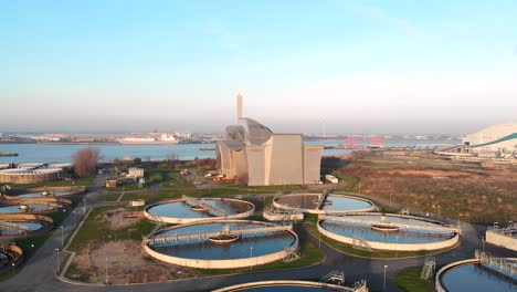 Aerial-Panning-Shot-around-Crossness-Sewage-Treatment-Works,-with-the-Thames-River-and-Boats-in-the-background