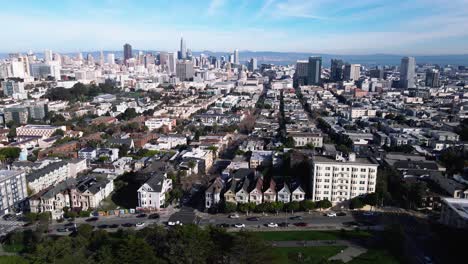 Drone-footage-presents-a-serene-view-of-San-Francisco's-Seven-Painted-Ladies,-iconic-Victorian-houses,-set-against-the-vibrant-cityscape-and-lush-greenery-of-Alamo-Square-Park