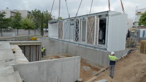 Construction-worker-guiding-modular-housing-unit-hanging-from-crane-into-foundation-position