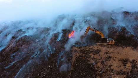 Vista-Aérea-De-Los-Bomberos-Usando-Una-Excavadora-Para-Extinguir-Un-Incendio-Forestal,-Humo-Azul-Elevándose-Sobre-La-Naturaleza-Dañada,-Noche-Nublada,-En-República-Dominicana---Dolly,-Disparo-De-Drone