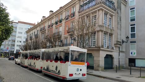 tourists-on-the-tram-passing-through-the-historic-pilgrimage-city,-nice-buildings-on-the-street-with-trees-and-people-strolling,-shot-blocked,-Santiago-de-Compostela,-Galicia