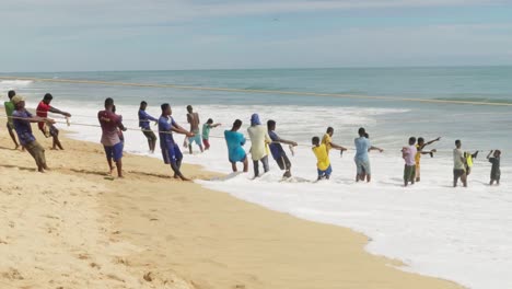 Hombres-Jóvenes-Tiran-Red-De-Pesca-Del-Océano-A-La-Playa-De-Arena-En-Ghana,-Amplia