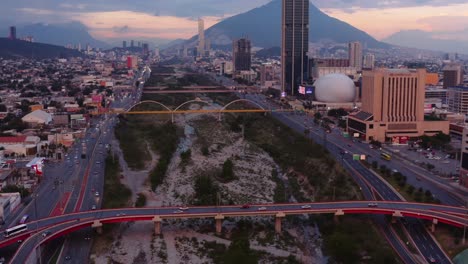 Die-Drohne,-Die-Bei-Sonnenuntergang-über-Den-Santa-Catarina-River-Aufsteigt,-Zeigt-Den-Belebten-Innenstadtbereich-Von-Monterrey,-Mexiko,-Mit-Hellen-Schildern-Und-Brücken,-Die-Das-Trockene-Flussbett-überqueren